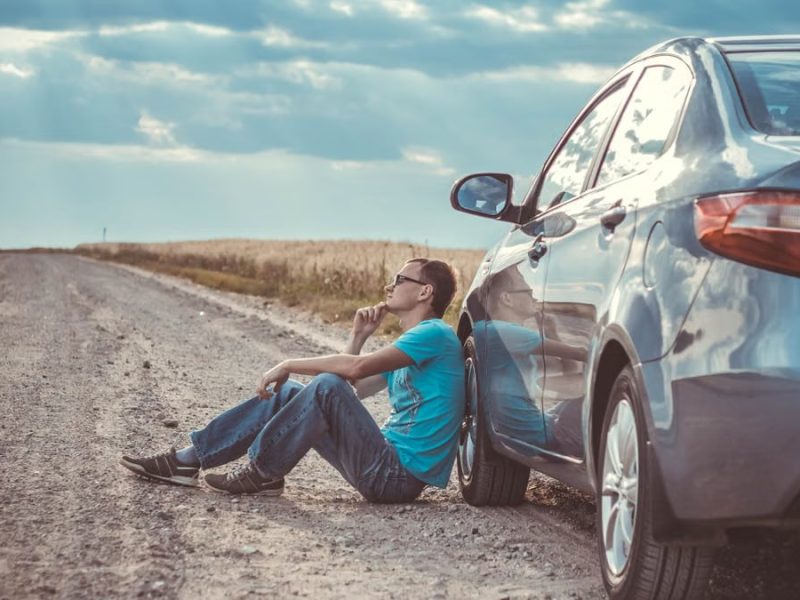 Man sitting by a car on a remote gravel road under a cloudy sky, deep in thought.